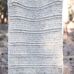K&ouml;nnte beinhalten: Eine gro&szlig;e, geh&auml;kelte Wolldecke mit einem strukturierten, gestreiften Muster in hellbeige. Die Decke hat Pompons an jeder Ecke.