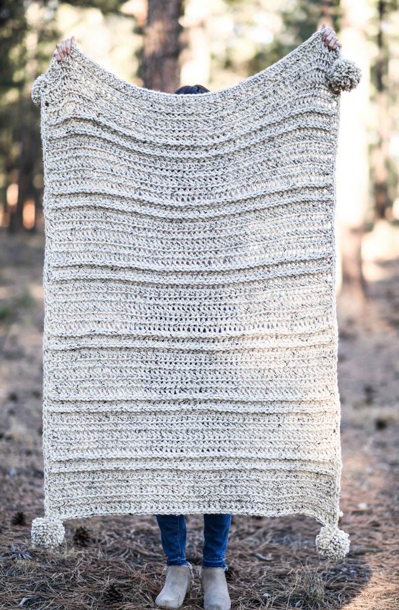 K&ouml;nnte beinhalten: Eine gro&szlig;e, geh&auml;kelte Wolldecke mit einem strukturierten, gestreiften Muster in hellbeige. Die Decke hat Pompons an jeder Ecke.
