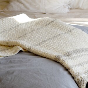 May include: A white and gray striped crocheted throw blanket on a gray bed.
