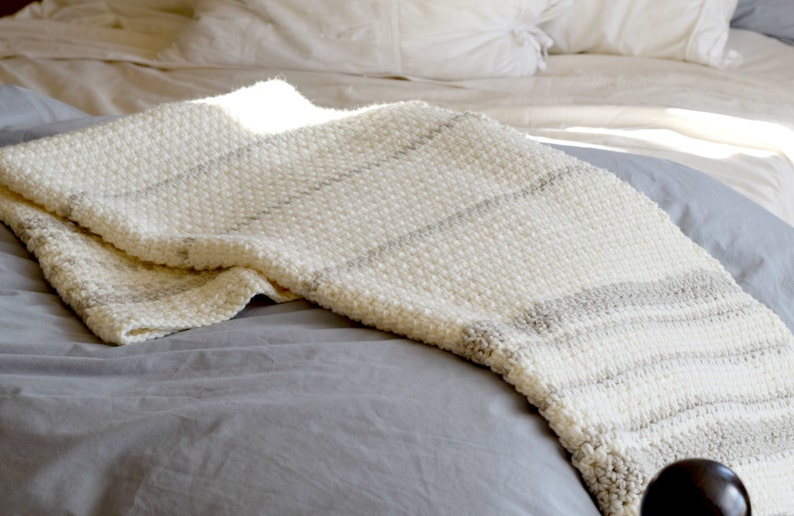 May include: A white and gray striped crocheted throw blanket on a gray bed.