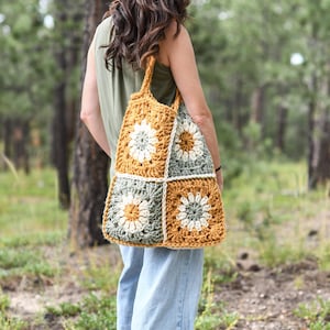 May include: A handmade crochet tote bag with a floral design. The bag features a patchwork pattern of mustard yellow, sage green, and white squares. The bag is carried over the shoulder and has a sturdy handle.