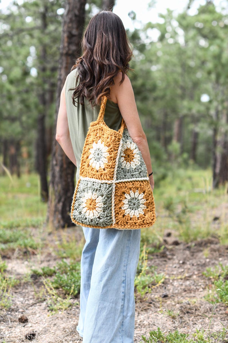 May include: A handmade crochet tote bag with a floral design. The bag features a patchwork pattern of mustard yellow, sage green, and white squares. The bag is carried over the shoulder and has a sturdy handle.