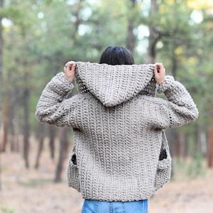 May include: A hooded crocheted cardigan in a light brown color with pockets. The cardigan is worn by a person in a forest setting.