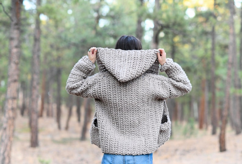 May include: A hooded crocheted cardigan in a light brown color with pockets. The cardigan is worn by a person in a forest setting.
