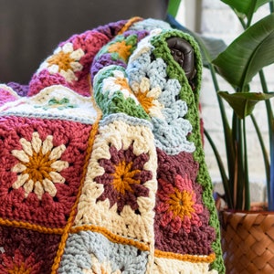 Könnte beinhalten: Eine gehäkelte Wolldecke mit Patchwork-Muster in verschiedenen Brauntönen, Grün, Blau und Creme. Die Decke besteht aus Quadraten mit einem Blumenmotiv in der Mitte.