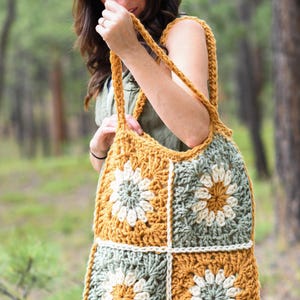 May include: A handmade crochet tote bag featuring a floral design. The bag is made with mustard yellow, sage green, and cream-colored yarn. The bag has a square shape with a large handle.