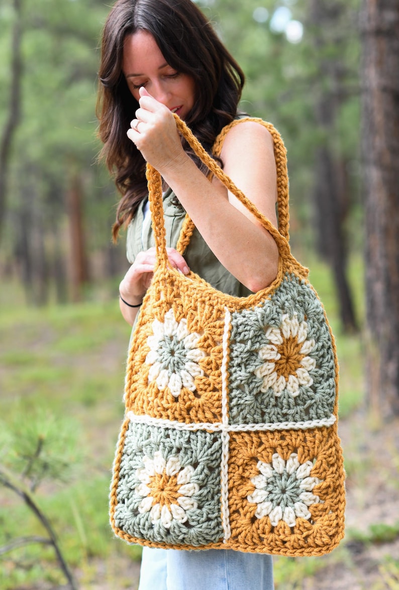 May include: A handmade crochet tote bag featuring a floral design. The bag is made with mustard yellow, sage green, and cream-colored yarn. The bag has a square shape with a large handle.