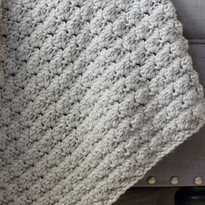 May include: A soft, grey crocheted throw blanket draped over a grey sofa. The blanket has a textured, raised stitch pattern.