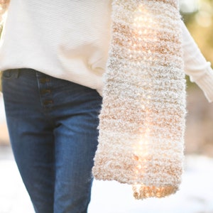 May include: A woman wearing a white sweater and blue jeans with a white and beige fuzzy scarf draped around her shoulders.