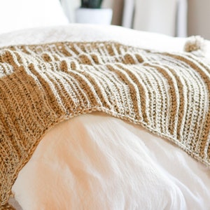 K&ouml;nnte beinhalten: Eine beige geh&auml;kelte Wolldecke mit Quasten an den Ecken, &uuml;ber ein wei&szlig;es Bett gelegt.