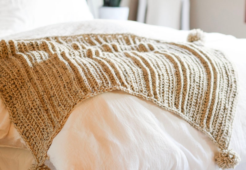 K&ouml;nnte beinhalten: Eine beige geh&auml;kelte Wolldecke mit Quasten an den Ecken, &uuml;ber ein wei&szlig;es Bett gelegt.