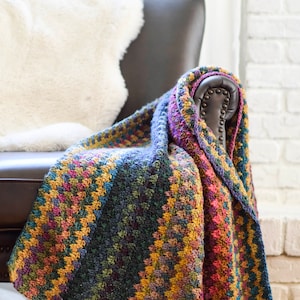 Pu&ograve; includere: Una coperta in crochet multicolore drappeggiata su una poltrona in pelle. La coperta presenta un motivo a chevron in tonalit&agrave; di viola, rosa, blu, verde e giallo.