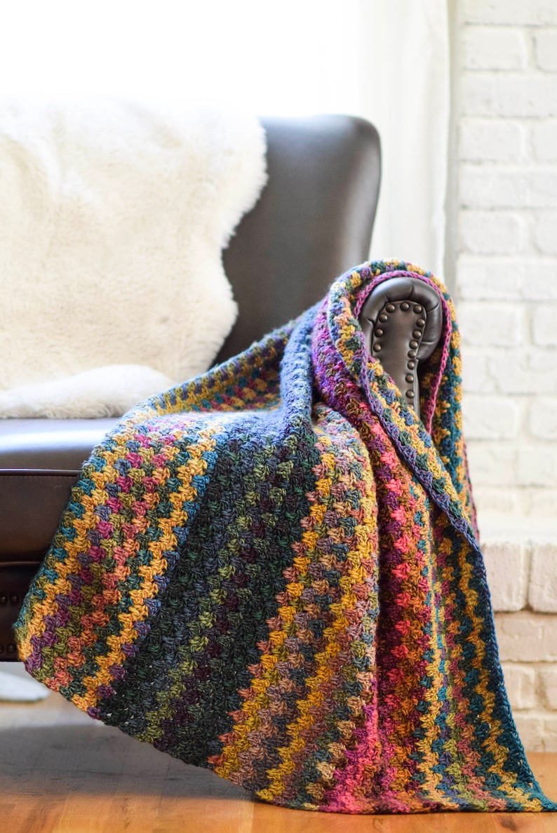 Pu&ograve; includere: Una coperta in crochet multicolore drappeggiata su una poltrona in pelle. La coperta presenta un motivo a chevron in tonalit&agrave; di viola, rosa, blu, verde e giallo.