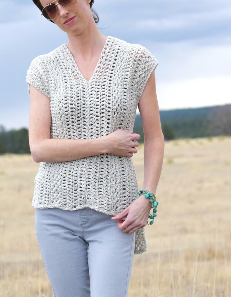 Puede incluir: Un top de crochet con cuello en V y mangas cortas. El top es de color beige claro y tiene un corte holgado y fluido. La modelo lleva el top con pantalones vaqueros grises claros.