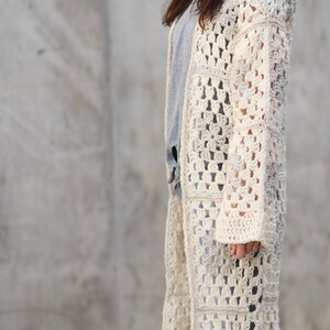 May include: A long, off-white crochet cardigan with a square stitch pattern. The cardigan has long sleeves and a loose fit.