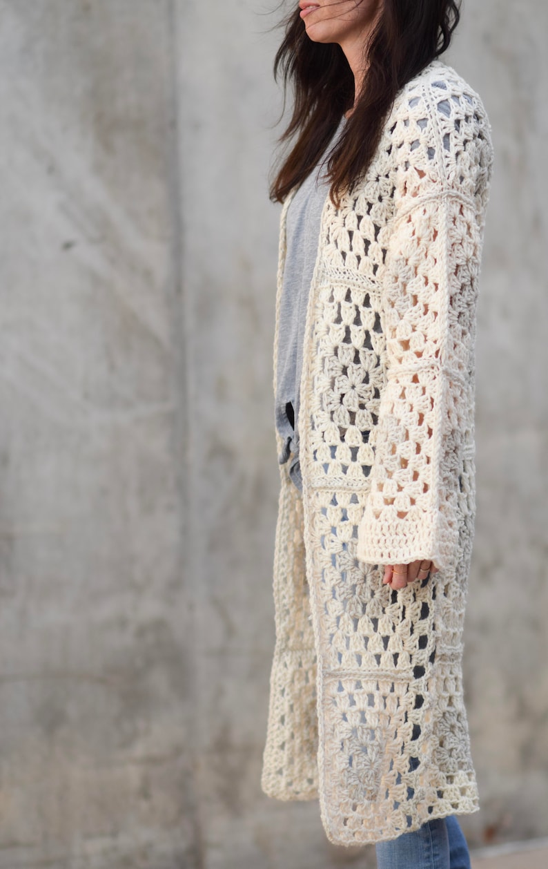 May include: A long, off-white crochet cardigan with a square stitch pattern. The cardigan has long sleeves and a loose fit.
