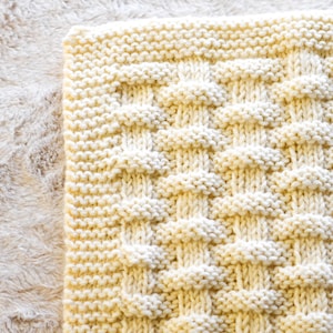May include: Close-up of a cream-coloured knitted blanket with a textured, raised pattern. The blanket is rectangular and appears soft and cosy. The knitting pattern creates a grid-like design with raised, rounded elements.