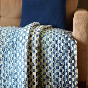 May include: A blue, green and white crocheted throw blanket folded on a brown sofa with a blue cushion.