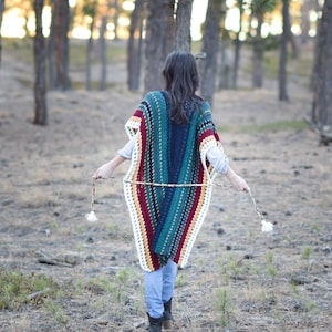 May include: A woman wearing a colorful striped crochet poncho with white fringe. The poncho has a deep blue, teal, green, yellow, red, and white color scheme. The woman is standing in a forest setting.