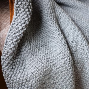 Puede incluir: Primer plano de una suave manta de punto gris, drapeada sobre una silla de madera. La manta tiene un aspecto texturizado y tejido con un borde grueso. El color es un gris apagado y neutro.