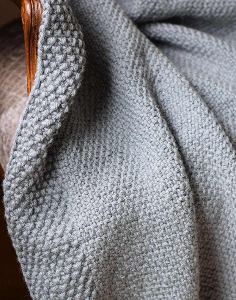 Puede incluir: Primer plano de una suave manta de punto gris, drapeada sobre una silla de madera. La manta tiene un aspecto texturizado y tejido con un borde grueso. El color es un gris apagado y neutro.