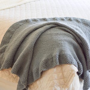 Puede incluir: Una manta gris tejida con un borde texturizado descansa sobre una cama. La manta est&aacute; sobre un edred&oacute;n beige acolchado y una colcha blanca acolchada. La textura suave y el color neutro crean una est&eacute;tica acogedora.