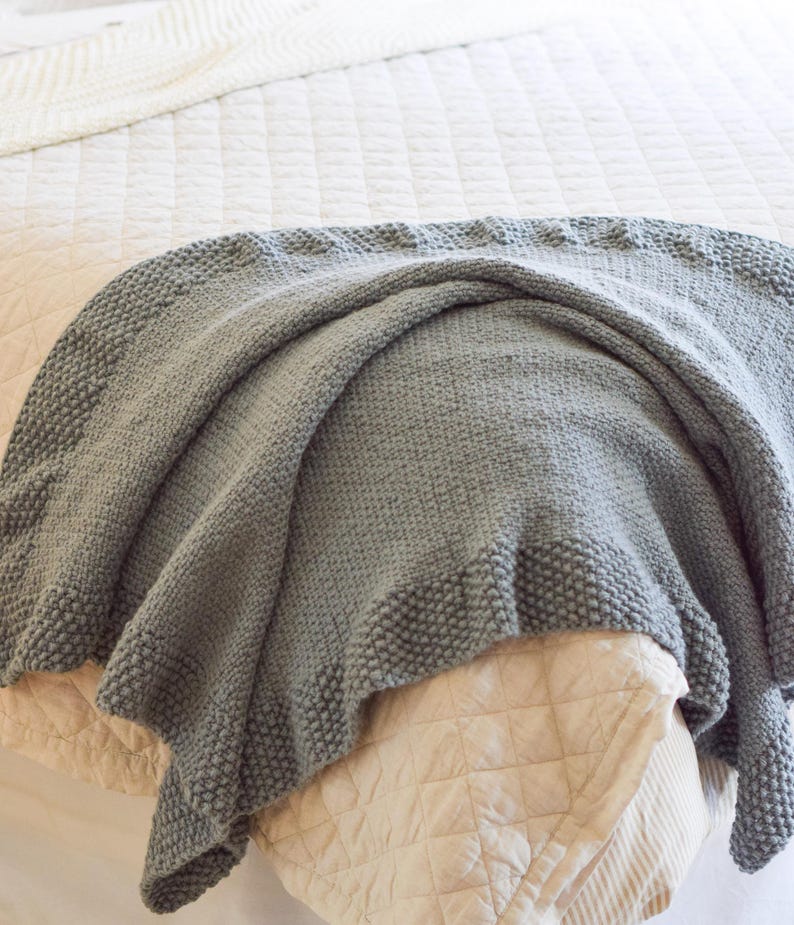 Puede incluir: Una manta gris tejida con un borde texturizado descansa sobre una cama. La manta est&aacute; sobre un edred&oacute;n beige acolchado y una colcha blanca acolchada. La textura suave y el color neutro crean una est&eacute;tica acogedora.