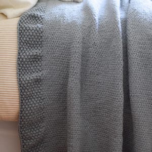 Puede incluir: Una manta de punto gris suave con una superficie texturizada. La manta tiene un borde gris m&aacute;s oscuro y est&aacute; sobre una cama. Una almohada blanca con borlas es visible en el fondo.