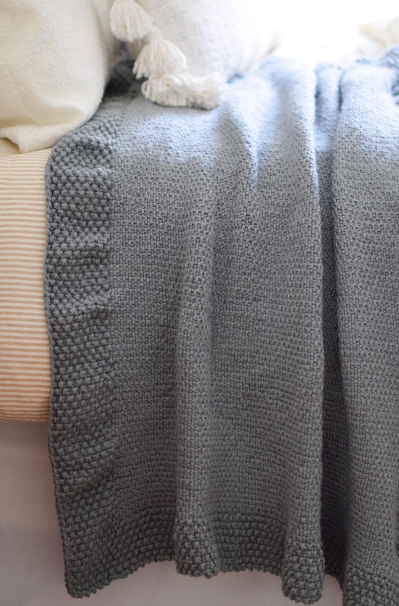 Puede incluir: Una manta de punto gris suave con una superficie texturizada. La manta tiene un borde gris m&aacute;s oscuro y est&aacute; sobre una cama. Una almohada blanca con borlas es visible en el fondo.