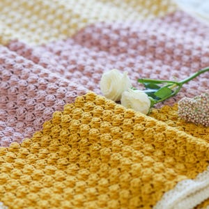 May include: A handmade crochet blanket in shades of yellow, pink, and cream. The blanket features a textured stitch pattern and a cream-colored border. Two white roses and a sprig of small white flowers are placed on the blanket.