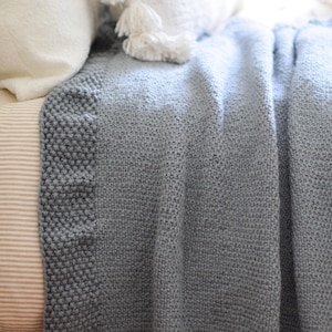 Puede incluir: Una manta de punto azul gris&aacute;ceo con un borde texturizado. La manta est&aacute; sobre una cama, con almohadas blancas y una almohada a rayas visibles en el fondo. El degradado de color de la manta a&ntilde;ade profundidad a la escena acogedora.