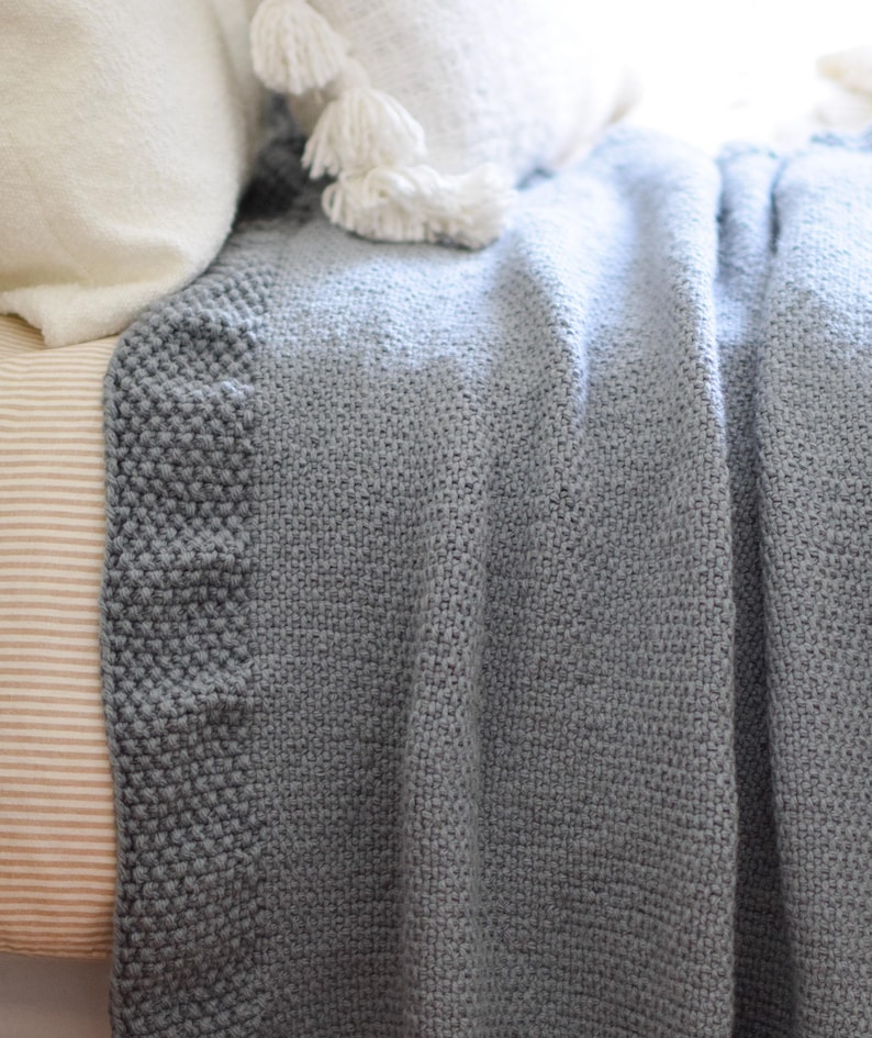 Puede incluir: Una manta de punto azul gris&aacute;ceo con un borde texturizado. La manta est&aacute; sobre una cama, con almohadas blancas y una almohada a rayas visibles en el fondo. El degradado de color de la manta a&ntilde;ade profundidad a la escena acogedora.