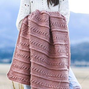 May include: A woman is holding a crocheted blanket in a dusty rose color. The blanket has a textured stitch pattern and is folded in half. The woman is wearing a white sweater and blue jeans.