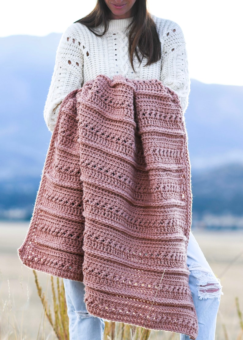 May include: A woman is holding a crocheted blanket in a dusty rose color. The blanket has a textured stitch pattern and is folded in half. The woman is wearing a white sweater and blue jeans.