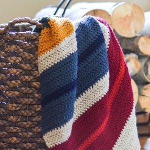May include: A striped crocheted blanket in shades of mustard yellow, navy blue, white, and burgundy red. The blanket is draped over a wicker basket.