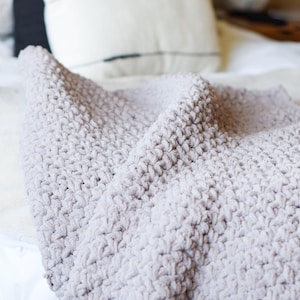 May include: A soft, gray, crocheted throw blanket with a textured, fluffy surface. The blanket is folded and draped over a bed with white bedding.