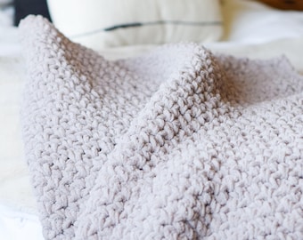 Könnte beinhalten: Eine weiche, graue, gehäkelte Wolldecke mit einer strukturierten, flauschigen Oberfläche. Die Decke ist gefaltet und über ein Bett mit weißen Bettwäsche drapiert.
