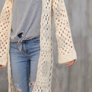 May include: A long, off-white crochet cardigan with a square stitch pattern. The cardigan is open front and has long sleeves. The model is wearing the cardigan over a grey t-shirt and blue jeans.