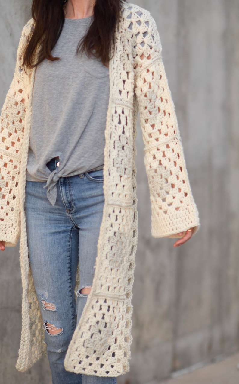 May include: A long, off-white crochet cardigan with a square stitch pattern. The cardigan is open front and has long sleeves. The model is wearing the cardigan over a grey t-shirt and blue jeans.