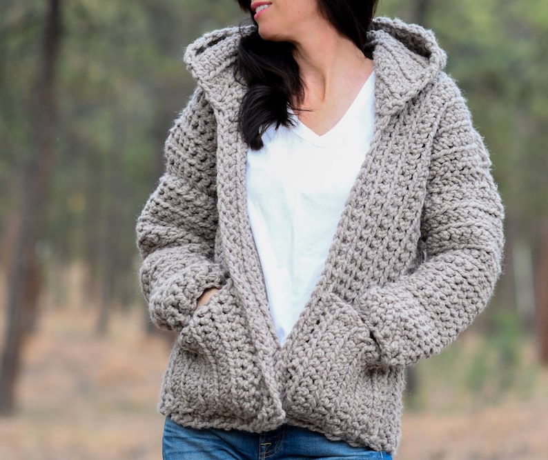 May include: A hooded cardigan with pockets in a light brown color. The cardigan is made of a soft, textured yarn.