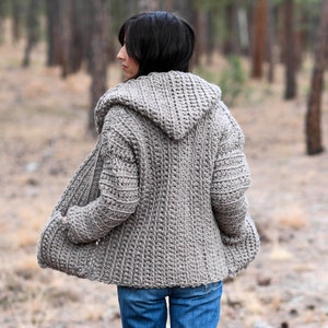 May include: A hooded crocheted cardigan in a light brown color. The cardigan is open in the front and has pockets on the sides. The person wearing the cardigan is standing in a forest.