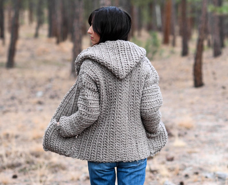 May include: A hooded crocheted cardigan in a light brown color. The cardigan is open in the front and has pockets on the sides. The person wearing the cardigan is standing in a forest.