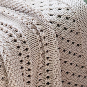 Peut inclure: Gros plan sur une couverture beige tricotée avec un motif texturé. La couverture présente une combinaison de tricot uni et de motifs ajourés, créant une surface visuellement intéressante et tactile. La couleur neutre convient à divers styles de décoration intérieure.