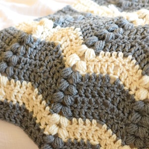 Może przedstawiać: Koc wykonany na szydełku z chevronowym wzorem w kolorze szarym i kremowym. Koc wykonany jest z fakturowanym ściegiem, który tworzy wypukły, puszysty efekt.