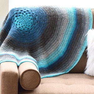 May include: A crocheted throw blanket in shades of blue and gray with a circular design. The blanket is draped over a brown couch with a white faux fur pillow.