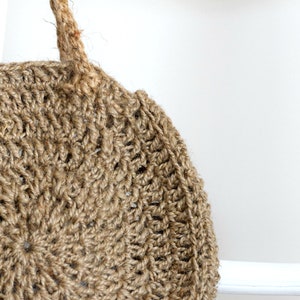 May include: A round, crocheted bag made of natural jute fiber with a brown color. The bag has two thick rope handles.