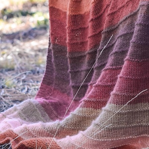 May include: A hand-knitted blanket in a gradient of warm colors, including shades of orange, pink, and brown. The blanket is draped over a surface, showcasing its textured, ribbed pattern. The soft, cozy blanket is perfect for adding warmth and style to any space.