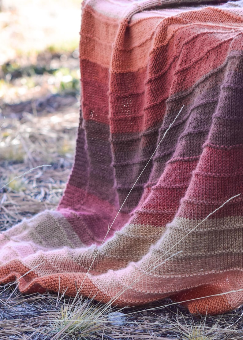 May include: A hand-knitted blanket in a gradient of warm colors, including shades of orange, pink, and brown. The blanket is draped over a surface, showcasing its textured, ribbed pattern. The soft, cozy blanket is perfect for adding warmth and style to any space.