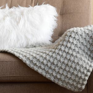 May include: A grey crocheted throw blanket with a textured pattern is draped over a brown sofa with a white faux fur cushion.