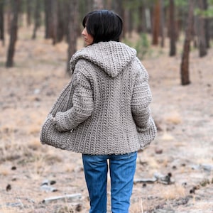May include: A hooded cardigan in a light brown color, worn by a person in a forest setting. The cardigan has a relaxed fit and a long, flowing style.